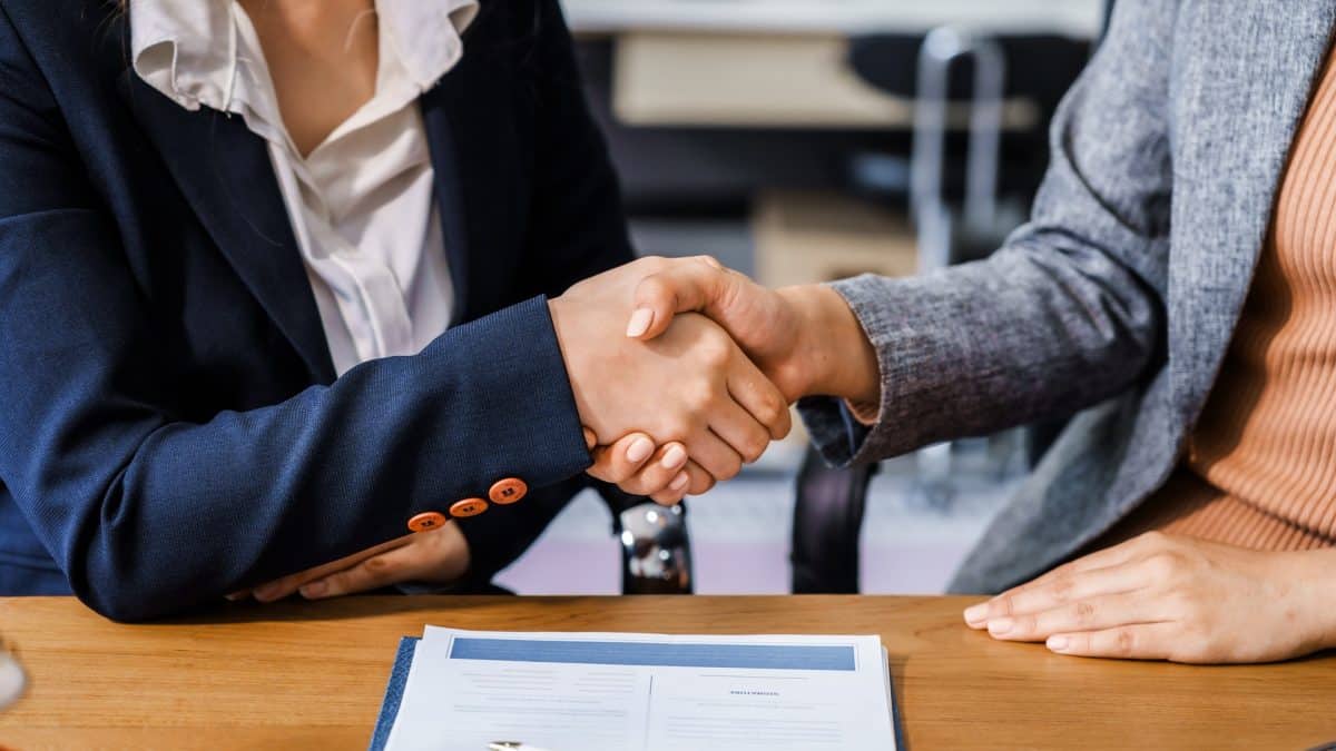 Two female lawyers in business suits shake hands at wooden desk, finalizing legal agreement. wooden gavel and scales represent justice as they discuss corporate law, compliance, dispute resolution.
