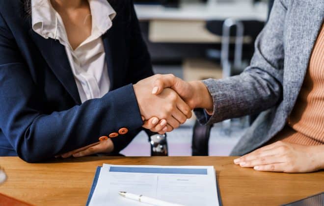 Two female lawyers in business suits shake hands at wooden desk, finalizing legal agreement. wooden gavel and scales represent justice as they discuss corporate law, compliance, dispute resolution.