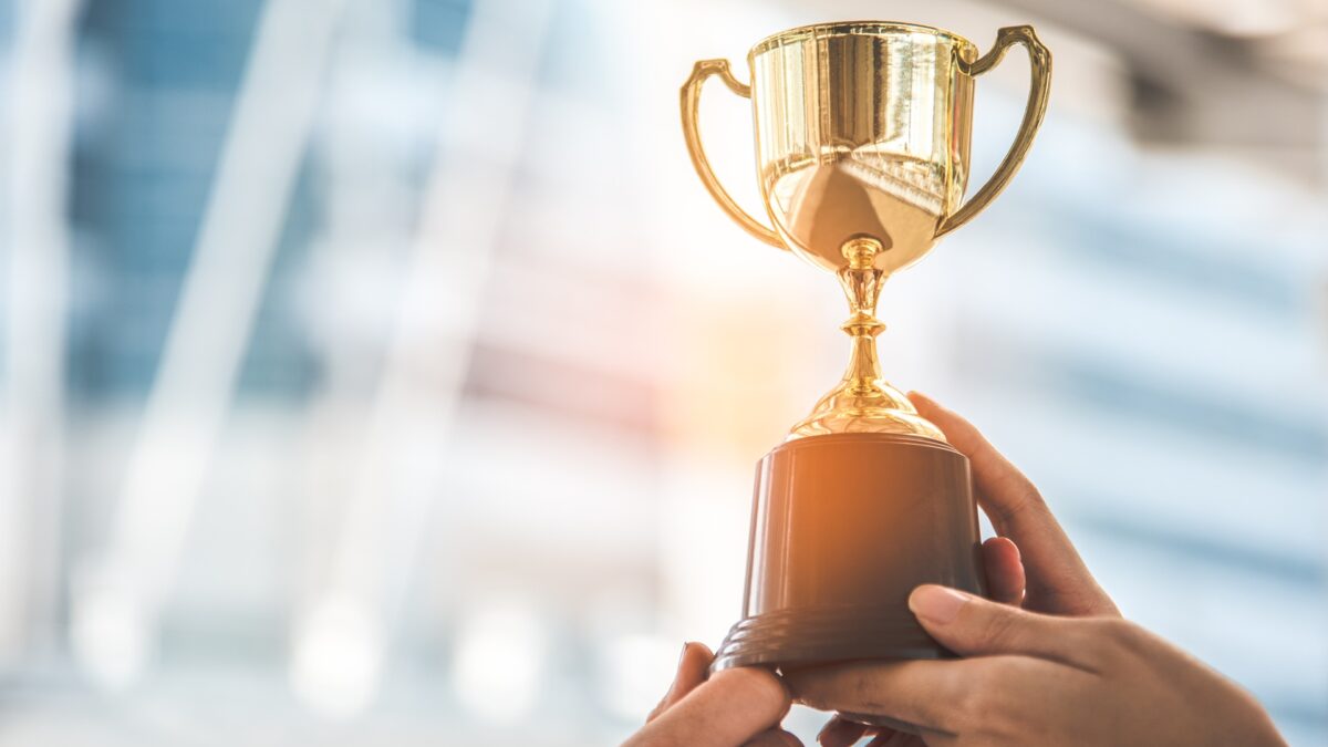 A golden trophy behind held in the air by two hands.