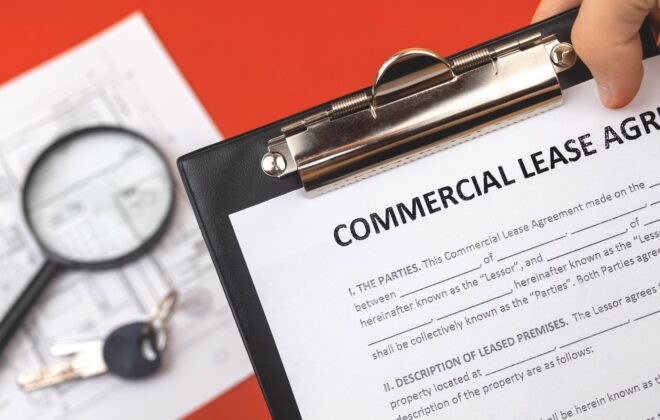 Hand holding a clipboard that has paperwork for a commercial lease agreement with a magnifying glass and keys in the background